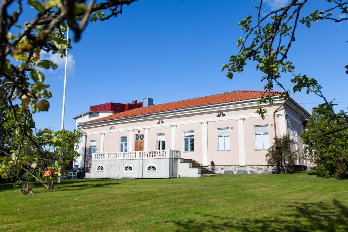 Kuvassa Opistotalo, rakennus on vaaleanpunainen kivitalo, jossa on oranssi laatta katto. Kuva on otettu kesällä ja edustalla on opistotalon puutarha, jossa on omenapuita ja vihreä nurmikko.